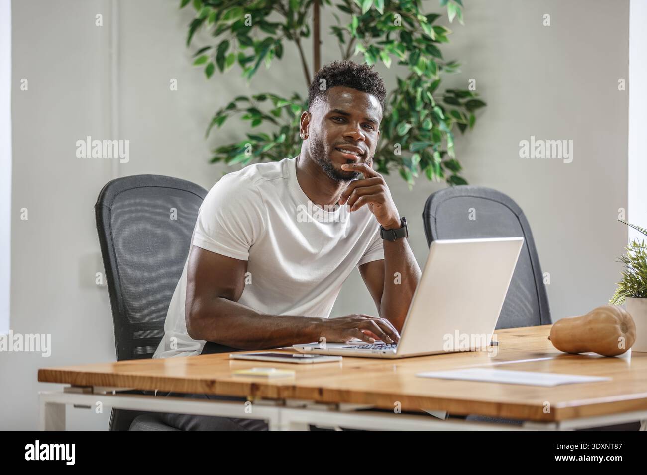 Un giovane uomo è concentrato sul proprio laptop, seduto su una scrivania di legno in un ufficio contemporaneo pieno di verde. Sembra rilassato e impegnato nel suo testamento Foto Stock