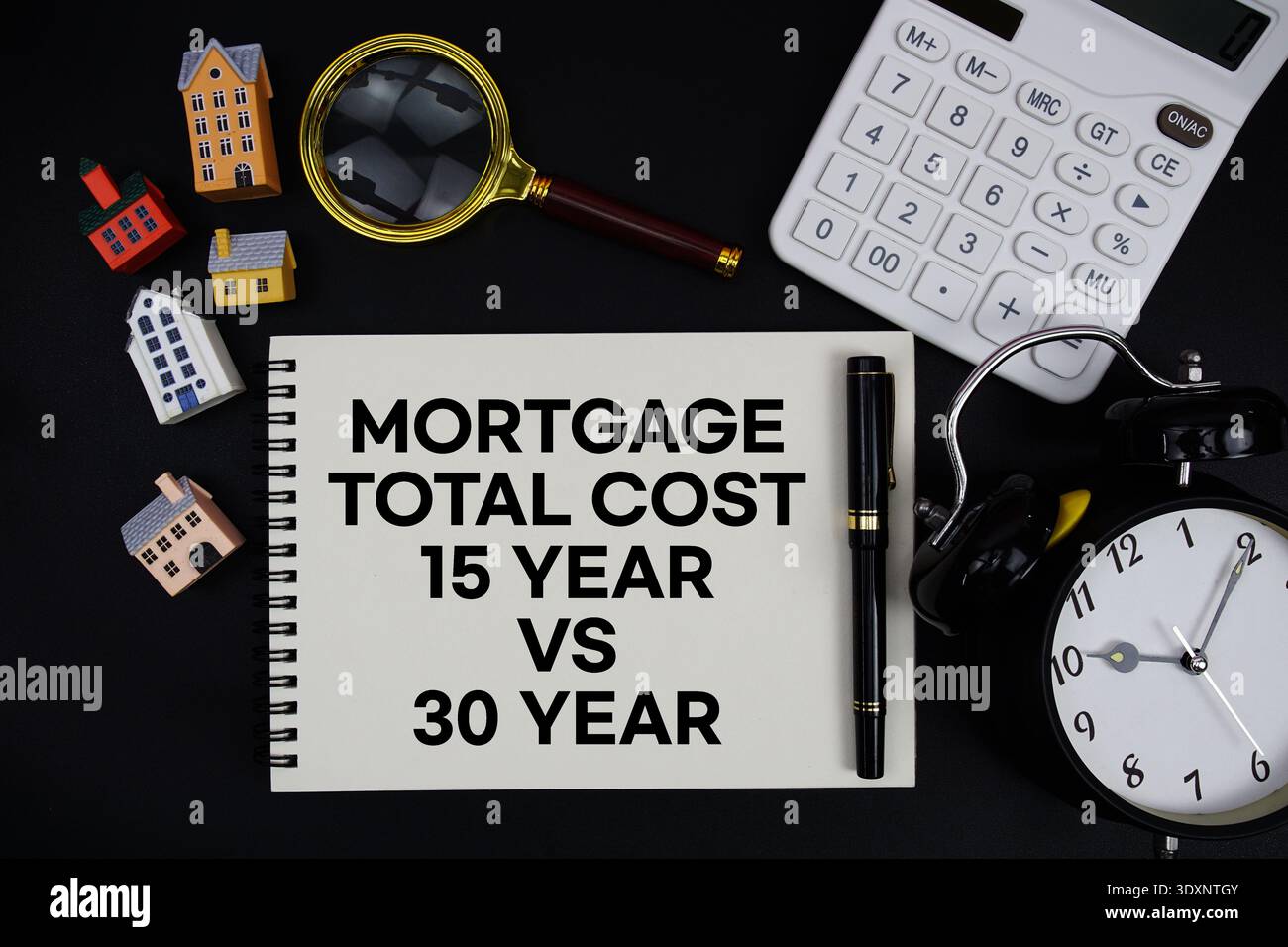 Blocco note con la frase "costo totale ipoteca 15 anni vs 30 anni" circondato da una penna, una calcolatrice, una lente di ingrandimento, un orologio e un modello di casa Foto Stock