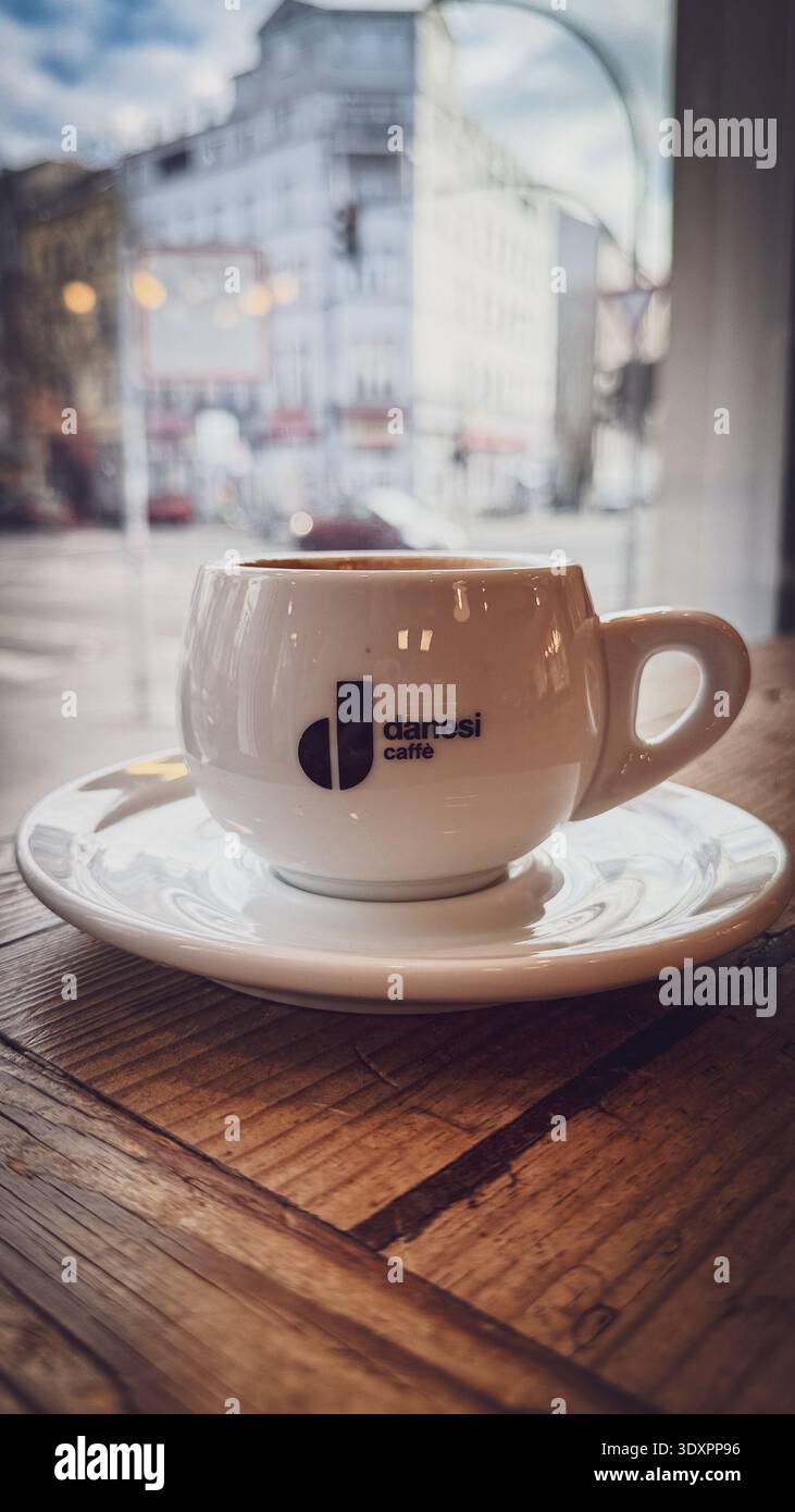 Tazza espresso danesi Caffe su piattino al tavolo di legno accanto alla finestra, vista sfocata della strada con edifici storici e cielo nuvoloso attraverso il bicchiere del caffè - Immagine stock catturata con smartphone