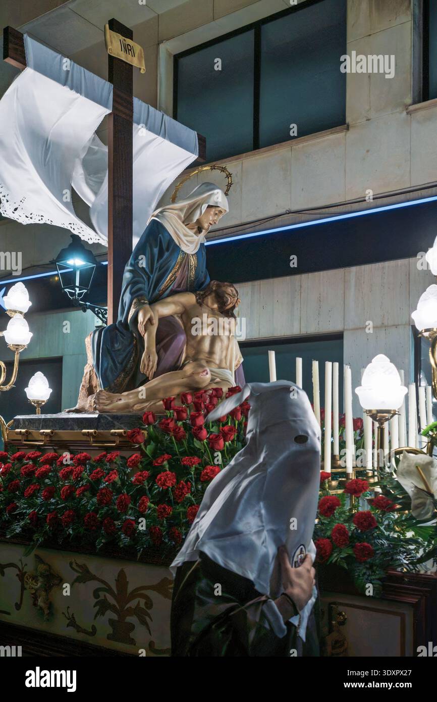 Processione della settimana Santa in Spagna con un galleggiante religioso Pietà adornato con rose rosse e un penitente in un tradizionale cappuccio bianco di notte. Foto Stock