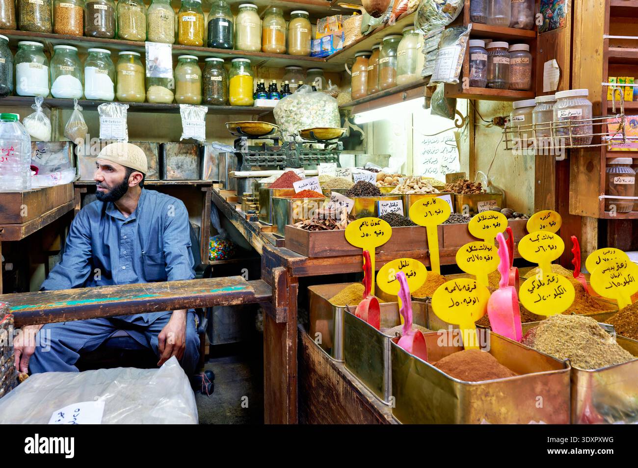 Un venditore siede tra barattoli e contenitori di spezie nel suq di al-Hamidiyah. I clienti navigano e fanno acquisti nell'affollato mercato di Damasco. Foto Stock