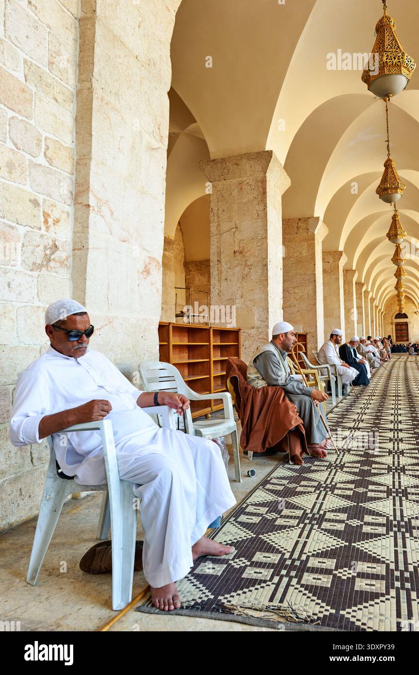 Gli uomini siedono tranquillamente nella moschea di Aleppo, in Siria, mentre aspettano. L'architettura mostra splendidi archi e muri in pietra. Foto Stock