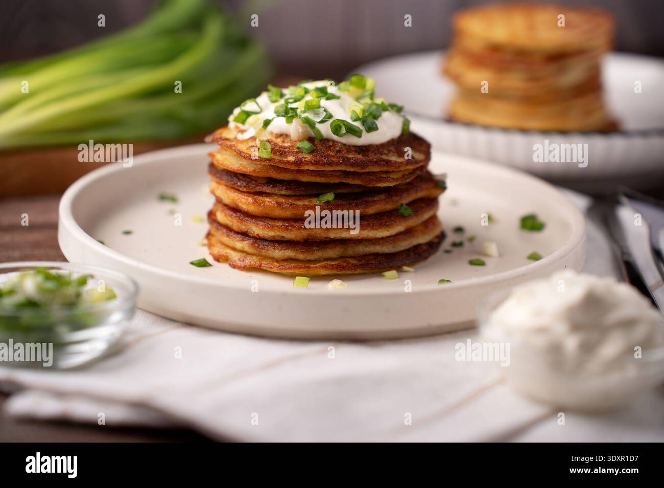 primo piano di un'alta pila di frittelle di patate dorate condite con crema all'arancia cremosa e erba cipollina tritata su un piatto di ceramica, un rustico tavolo in legno, morbido Foto Stock