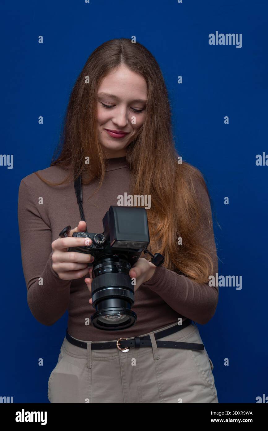 Giovane fotografa che rivede i controlli della fotocamera, tenendo la fotocamera digitale con un flash esterno e un obiettivo su sfondo blu Foto Stock