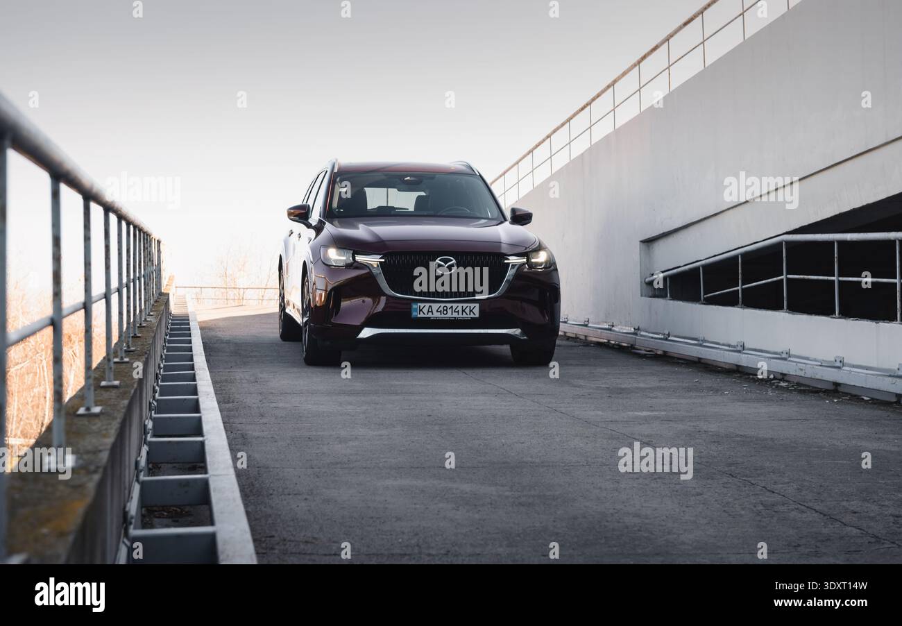 Vista frontale di una Mazda CX-90 rosso scuro su una rampa. SUV Mazda CX-90 Bordeaux scuro parcheggiato su una rampa di cemento, catturato dalla luce soffusa e chiara del giorno. Foto Stock