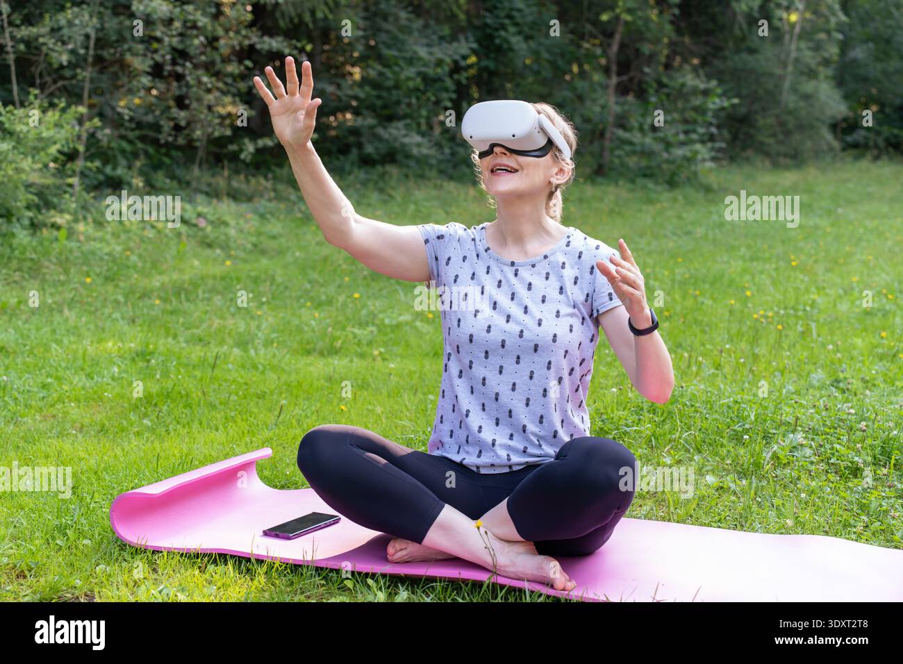 Una donna eccitata interagisce con un mondo di gioco virtuale mentre si siede su un tappetino yoga in natura. Foto Stock
