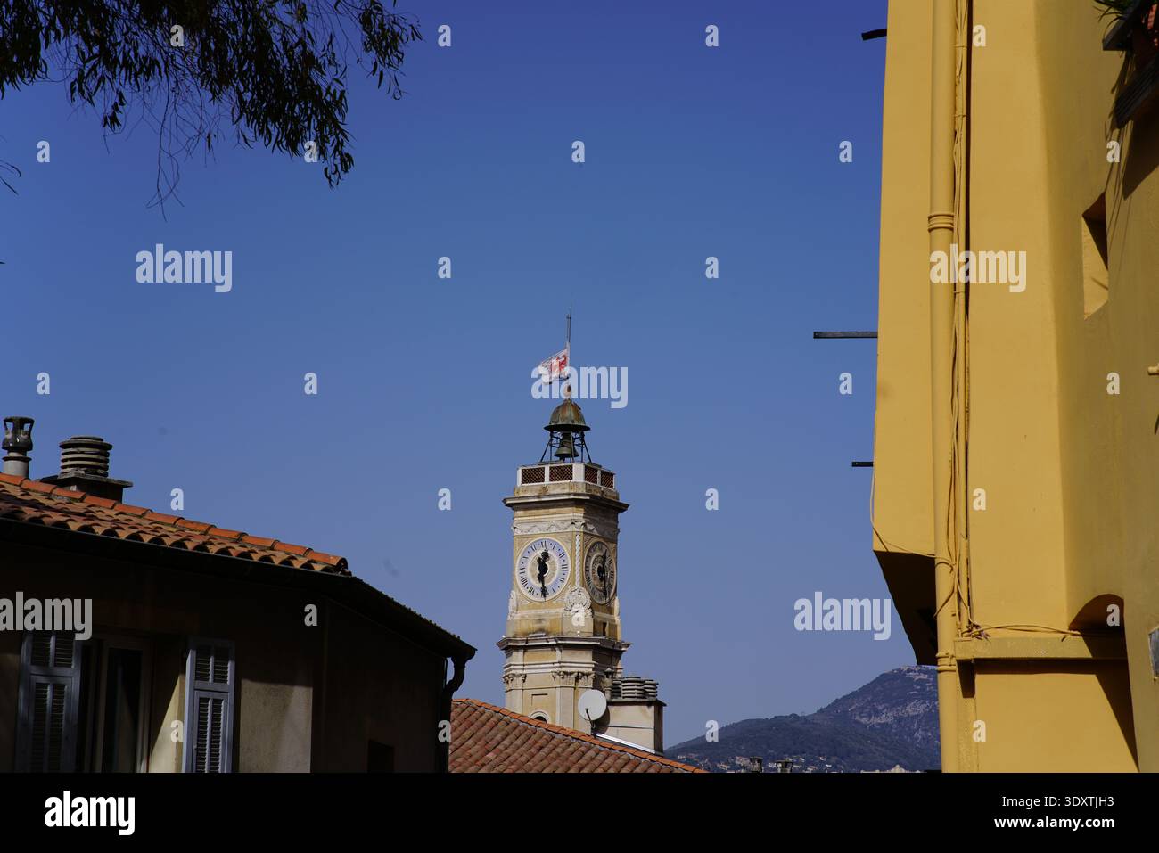 Storica torre dell'orologio con bandiera nella città vecchia di Vieux Nice incorniciata da edifici gialli sotto il cielo azzurro. Nizza, Francia Foto Stock