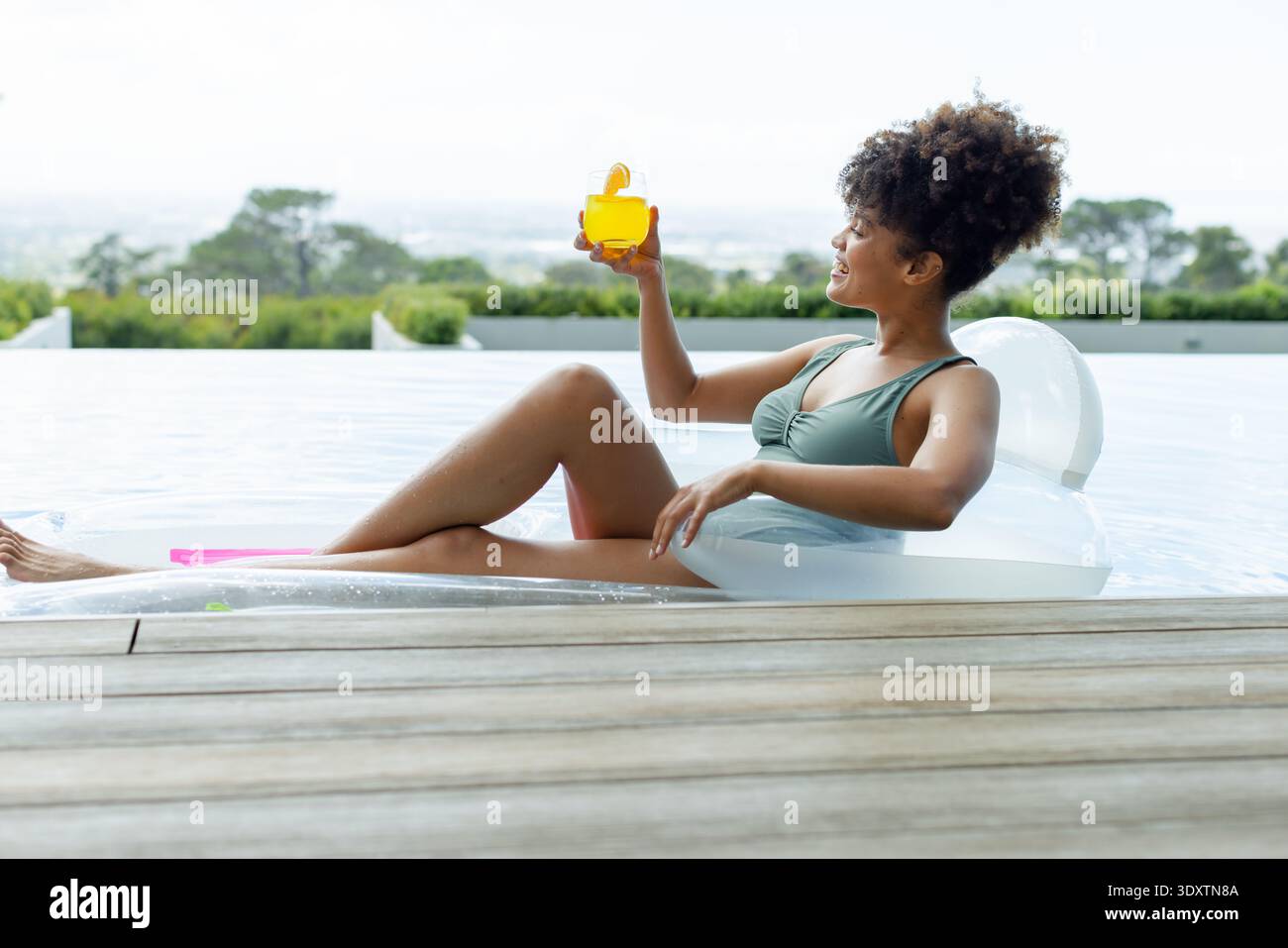 Il limpido salotto gonfiabile galleggia nella piscina a sfioro vicino al ponte in legno e scintillante bevanda gialla Foto Stock