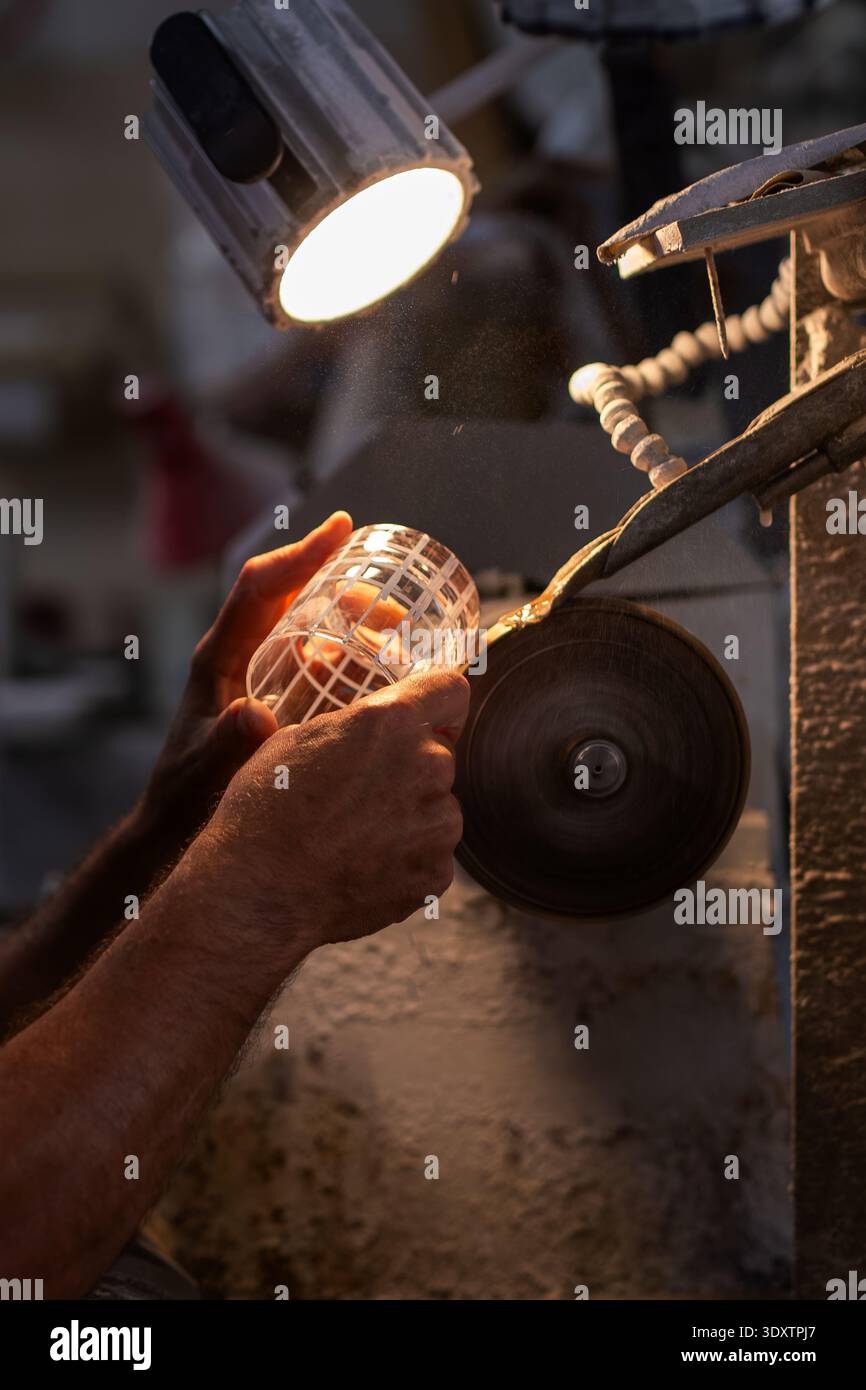 In un tradizionale laboratorio di Murano, un artigiano incide meticolosamente il vetro utilizzando una mola rotante. L'officina è poco illuminata, con una messa a fuoco Foto Stock