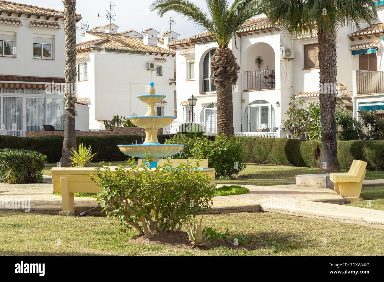 Un cortile soleggiato a Torrevieja, in Spagna, presenta una fontana colorata e palme, con edifici bianchi e mobili gialli, che creano un ambiente rilassante Foto Stock
