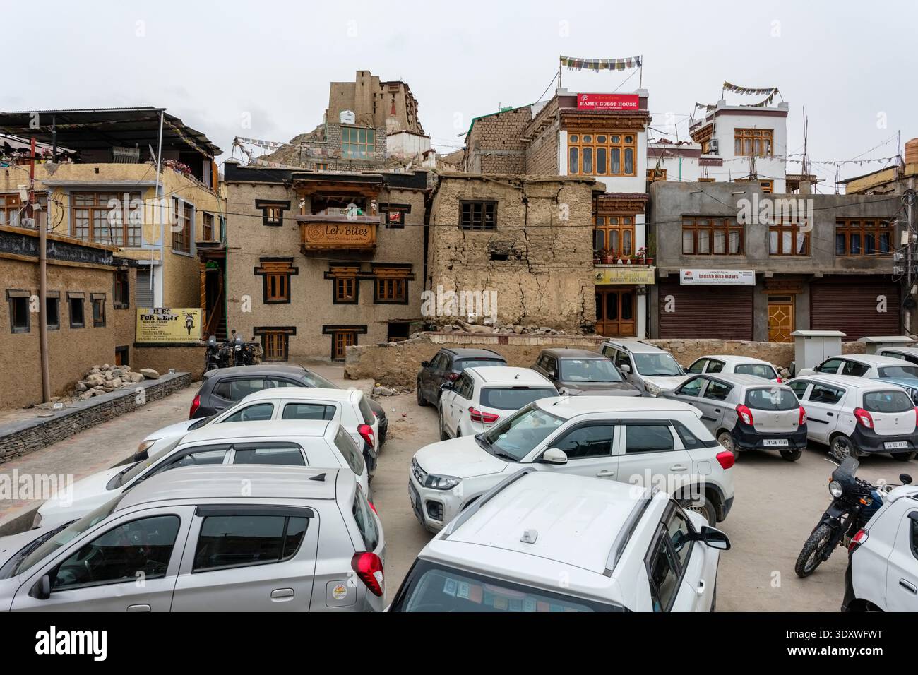 Edifici tradizionali a Leh con finestre intagliate e diverse auto in un parcheggio in primo piano, con Leh Palace in lontananza. Foto Stock