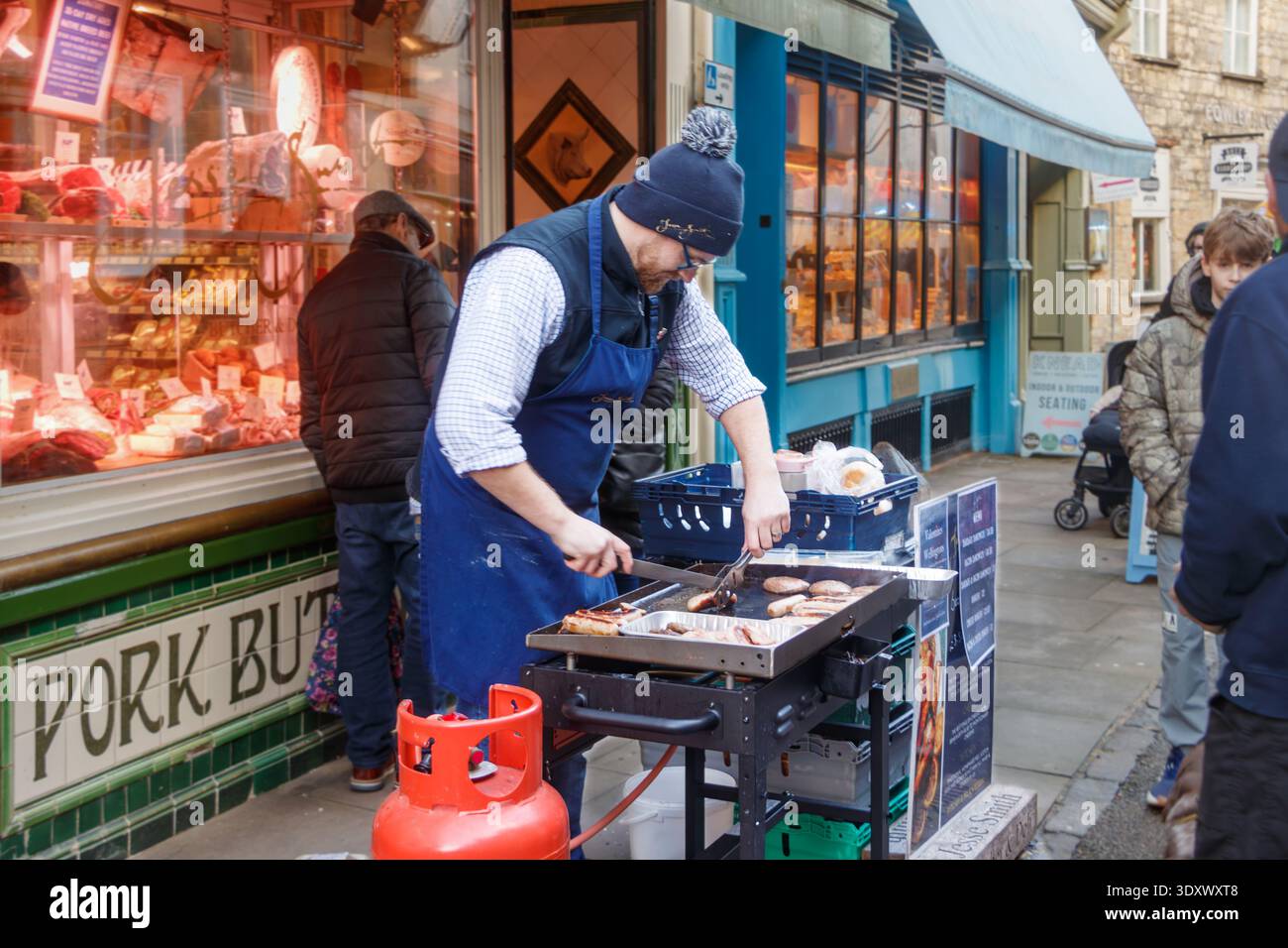 Il venditore ambulante cucina salsicce e hamburger su una piastra portatile fuori da una tradizionale macelleria in un ambiente urbano nel cirencester gloucestershire Foto Stock