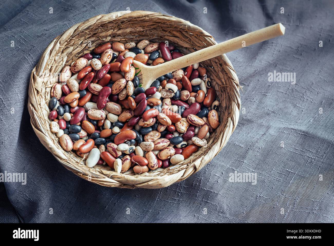 Varietà assortita di fagioli e legumi crudi essiccati in un cestello intrecciato su fondo rustico di stoffa scura. Foto Stock