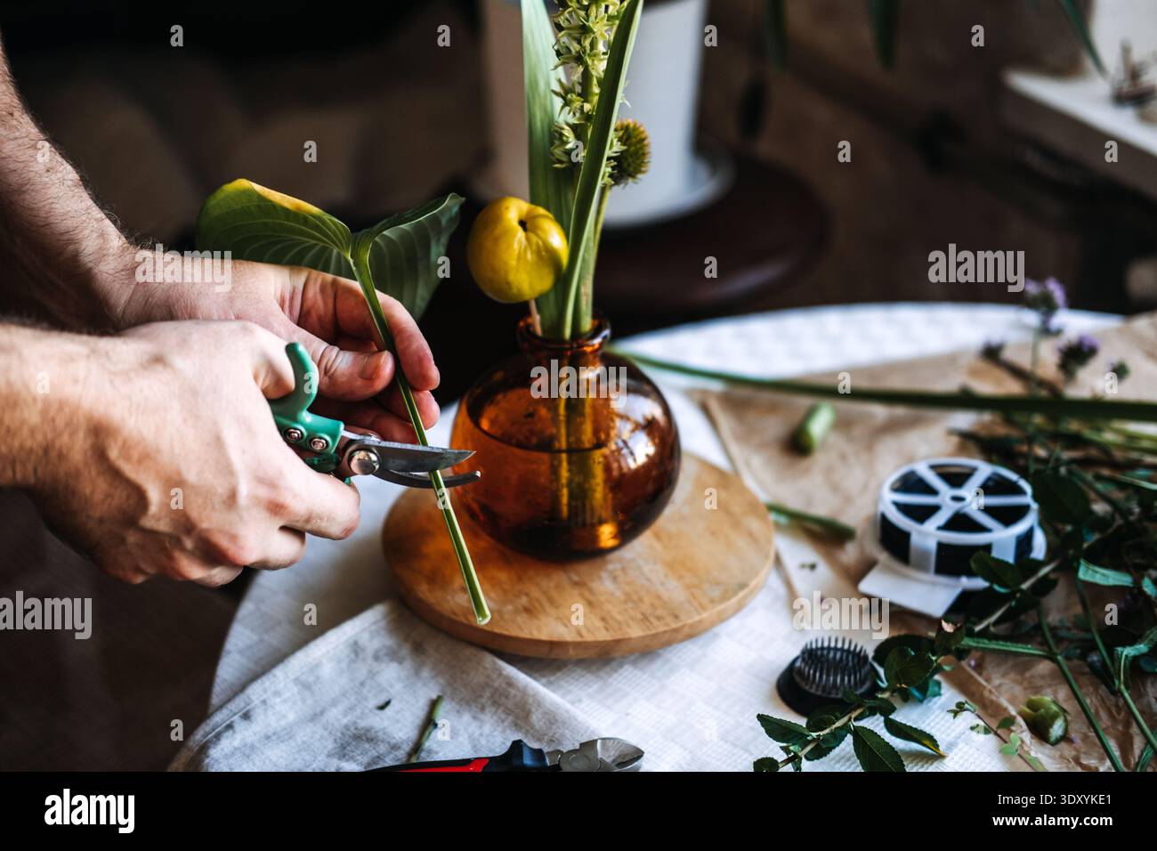 Le mani usano le cesoie da giardino per rifinire lo stelo verde vicino al vaso color ambra sul tavolo. Corso di progettazione floreale, formazione continua per creativi, formazione artigianale. Foto Stock