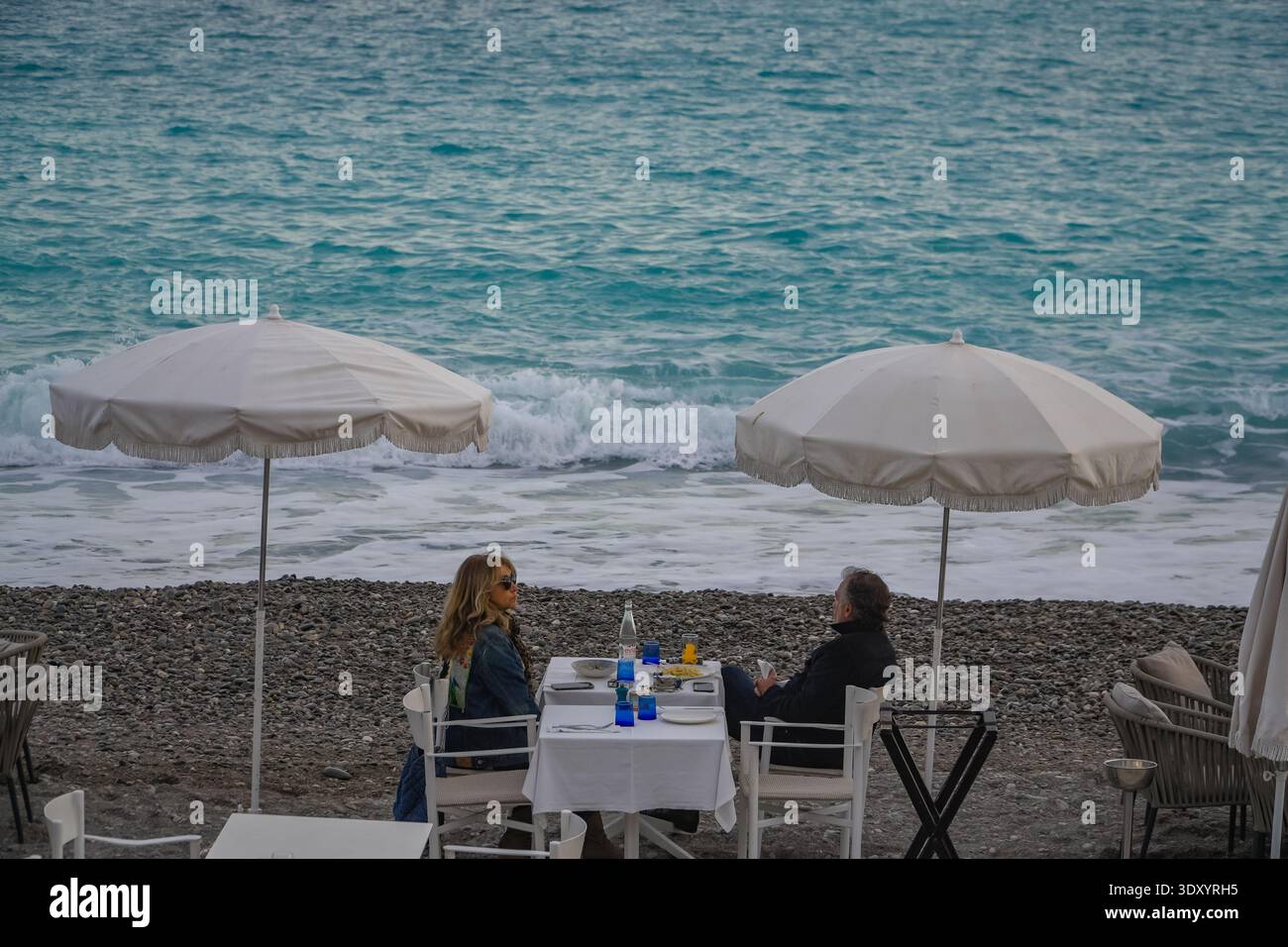 Ristoranti sulla spiaggia a Nizza, Francia. Coppia di mezza età che gusta un pasto sotto ombrelloni bianchi vicino alle onde turchesi del Mediterraneo. Foto Stock