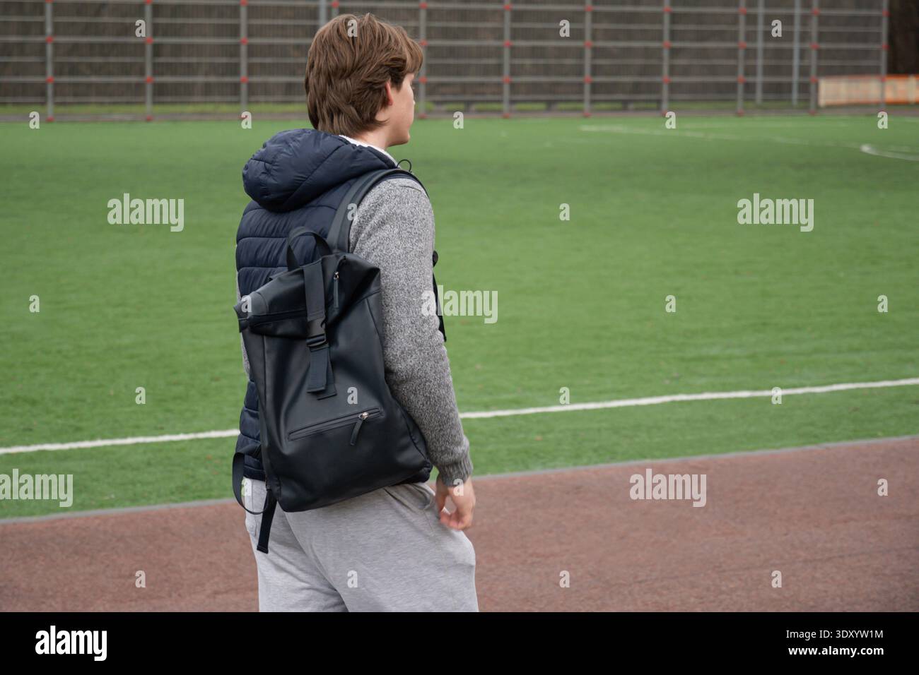 Camminando attraverso un campo sportivo all'aperto portando con sé uno zaino, un giovane uomo caucasico un giorno autunnale coperto dopo l'allenamento. Allenamento all'aperto Foto Stock