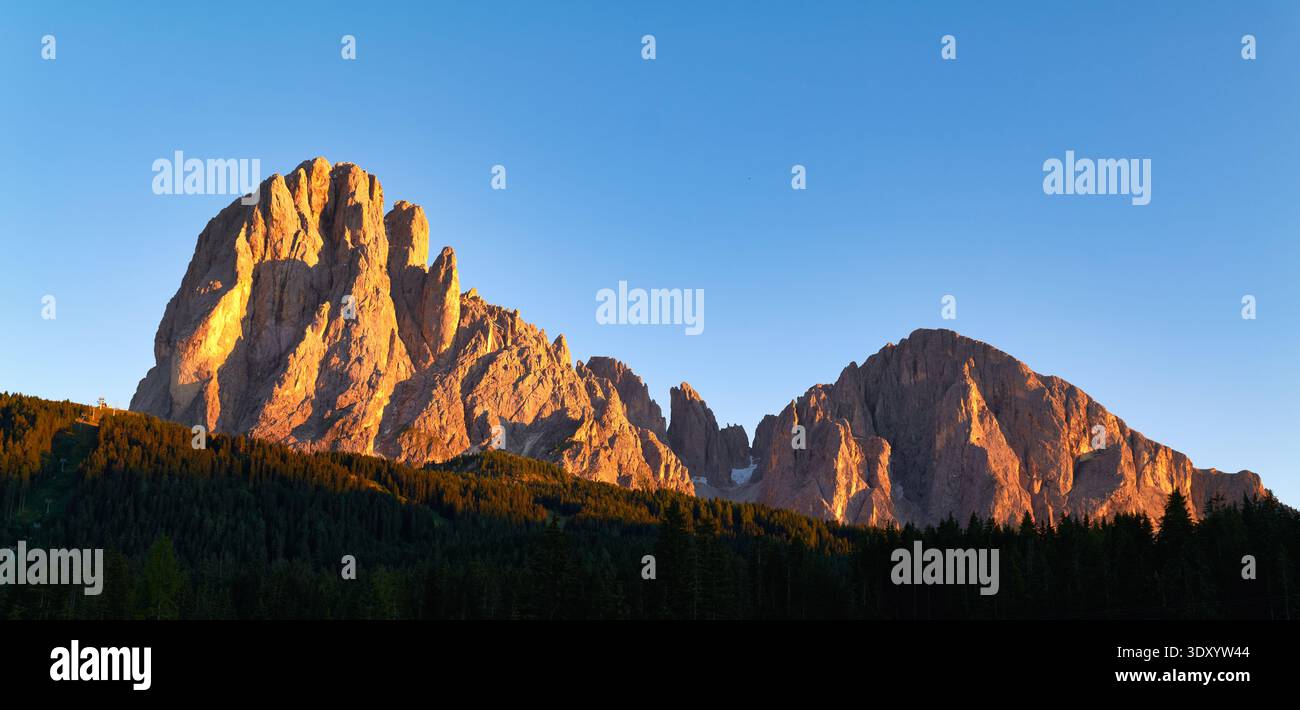 Il sole tramonta dietro il Sassolungo in Val Gardena, Italia. Le montagne brillano di luce arancione. Gli alberi riempiono il primo piano mentre il giorno diventa notte. Foto Stock