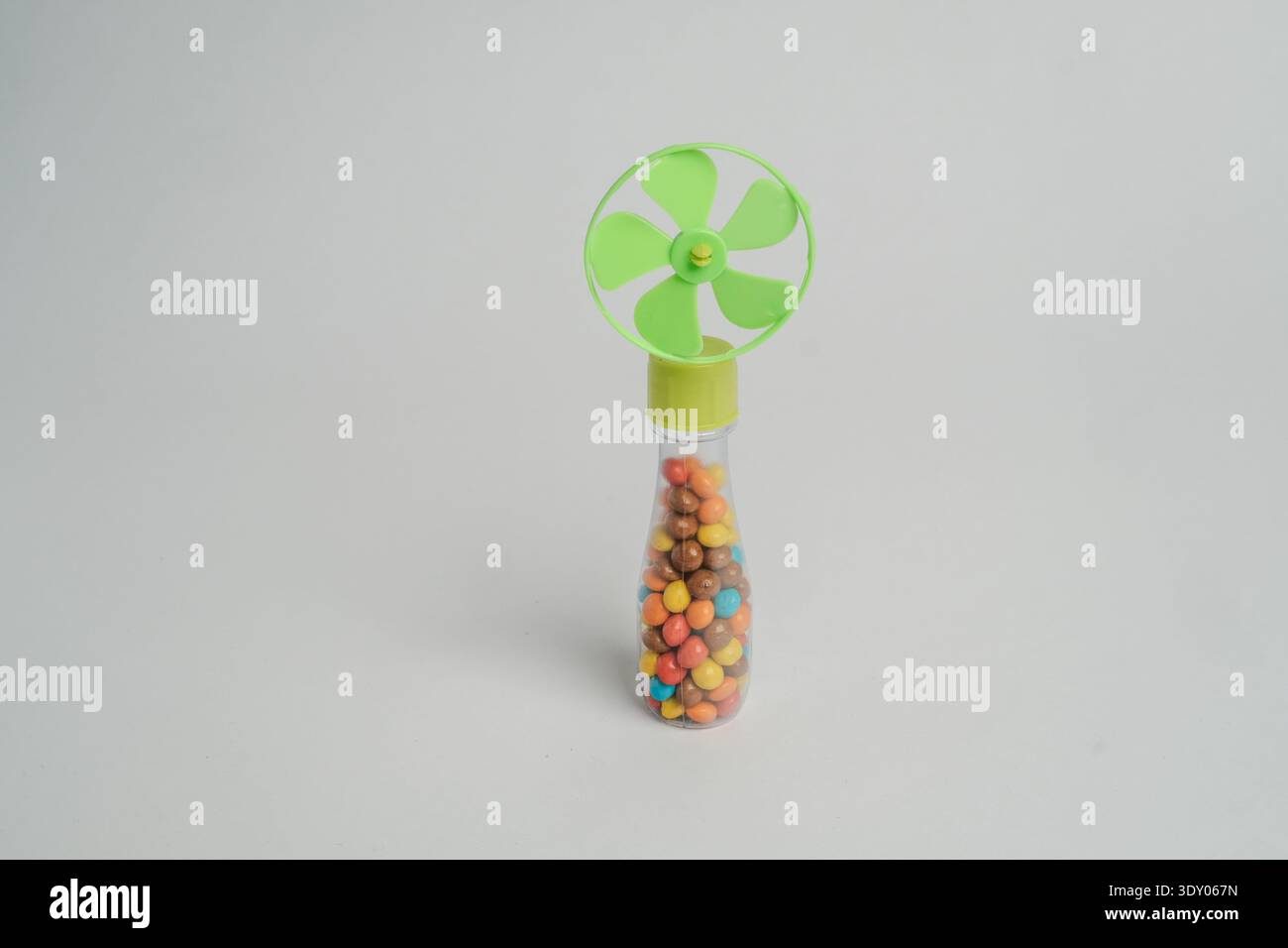 Piccolo ventilatore portatile in plastica per caramelle con parte superiore a elica verde e tubo trasparente riempito di caramelle colorate al cioccolato, isolato su sfondo grigio chiaro Foto Stock