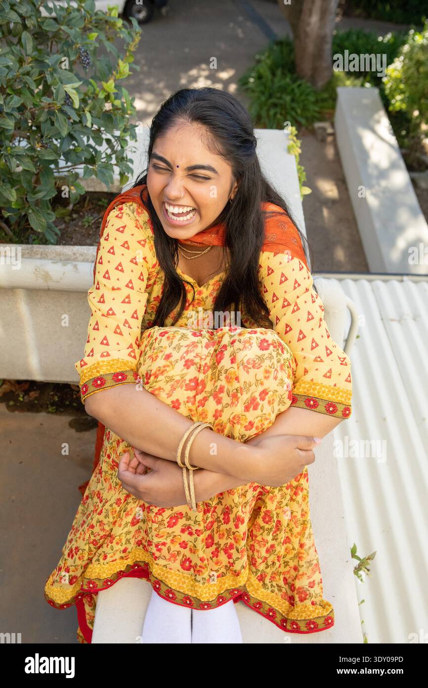 Donna indiana che abbraccia le ginocchia mentre ride su una panchina di pietra bianca sul terrazzo del giardino con dupatta rossa. Serenità, tempo libero, natura, tranquillità, cultura, vivace, outdoor || modello rilasciato Foto Stock