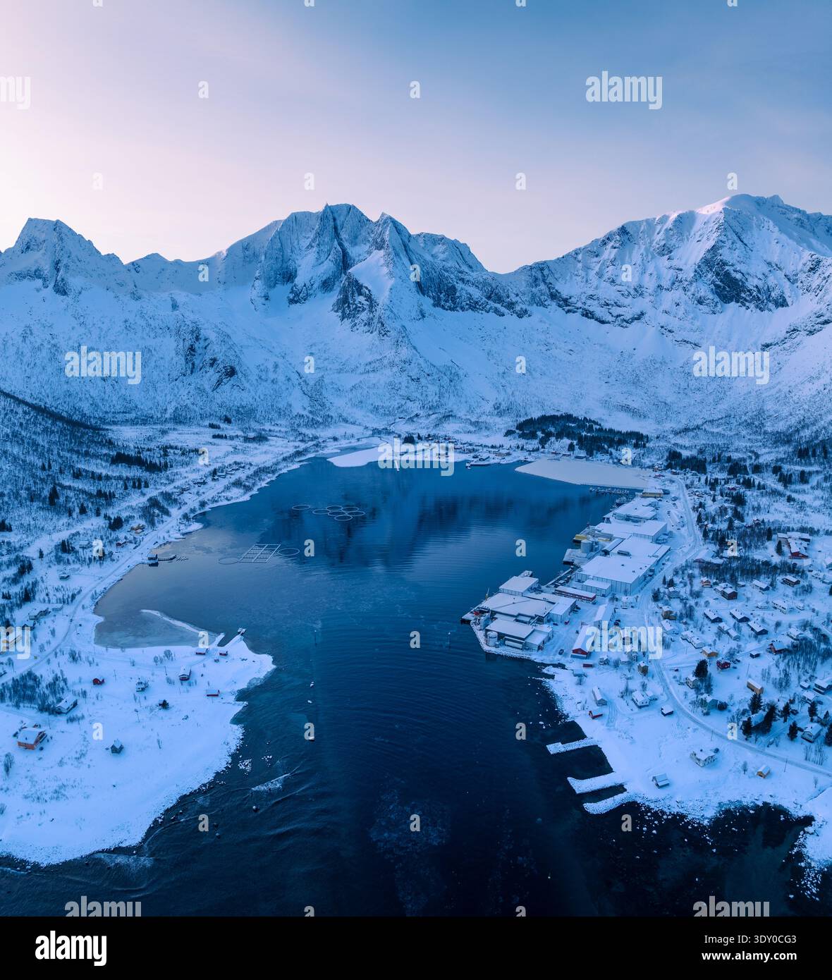 Vista aerea delle montagne innevate che si riflettono nelle tranquille acque di un fiordo, un tranquillo villaggio annidato lungo la costa, Senja, Norvegia. Foto Stock
