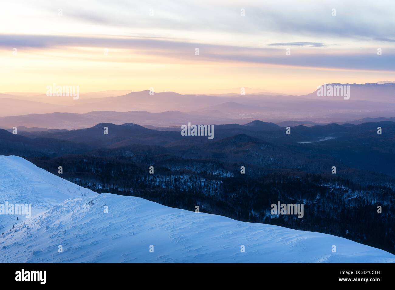 Vista del terreno innevato che incontra l'orizzonte con montagne in lontananza sotto un cielo pastello, Snežnik, Ilirska Bistrica, Slovenia. Foto Stock