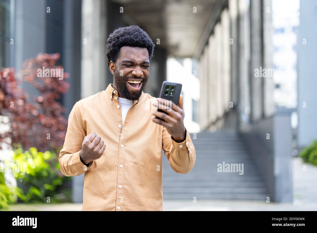 Persona felice che celebra il successo e la vittoria, guardando un telefono cellulare con un'espressione gioiosa e stringendo un pugno mentre si trova fuori da un edificio urbano Foto Stock