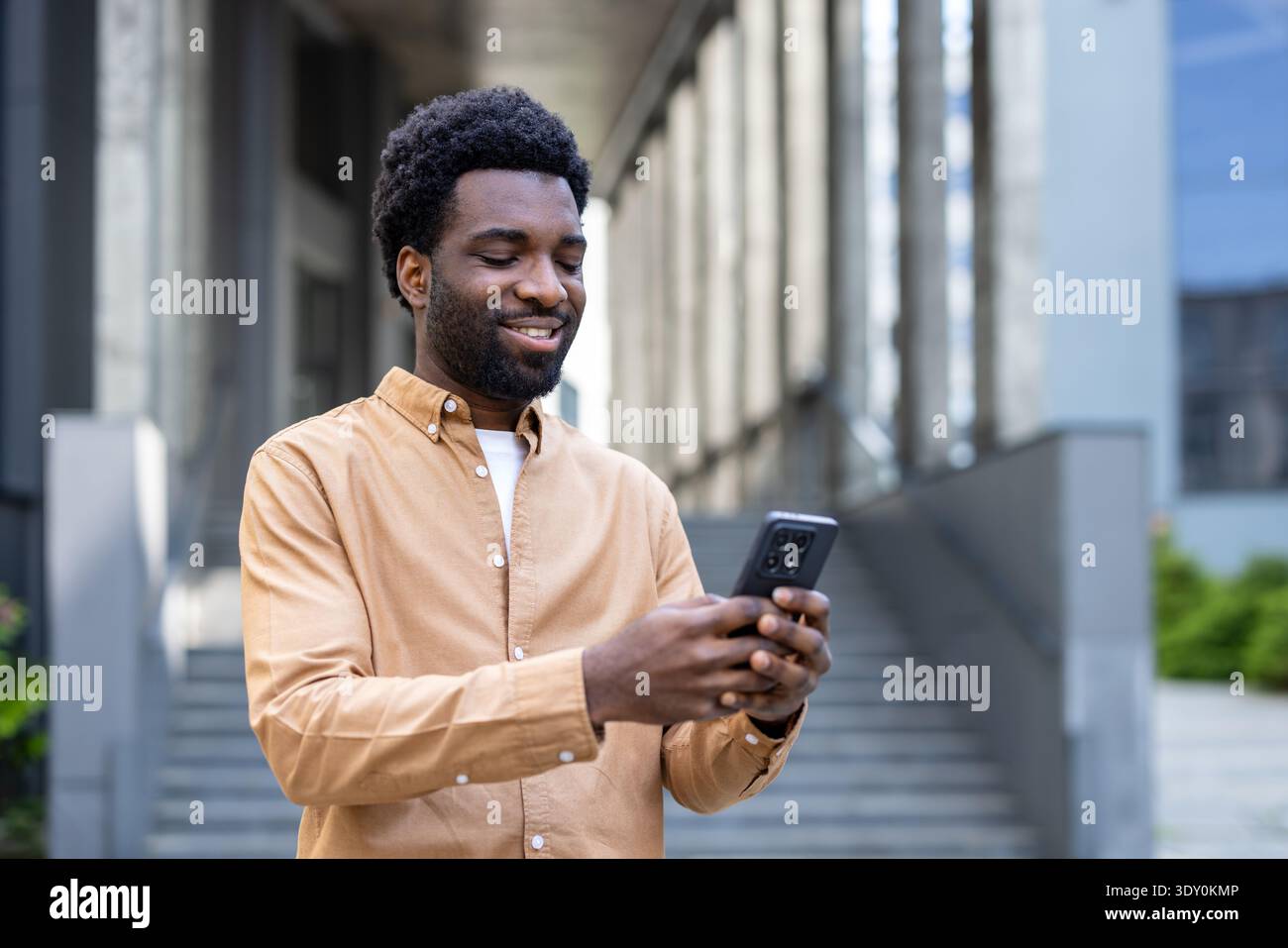 Giovane afroamericano in piedi su una strada urbana, sorridente e interagisce con il suo smartphone, rappresentando la comunicazione, il social network e lo stile di vita digitale moderno Foto Stock