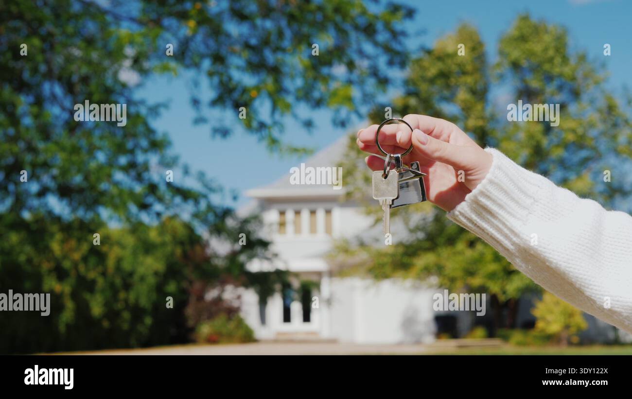 Una mano con un portachiavi a forma di piccola casa e chiavi. Sullo sfondo di una tipica casa in stile suburbano americano Foto Stock