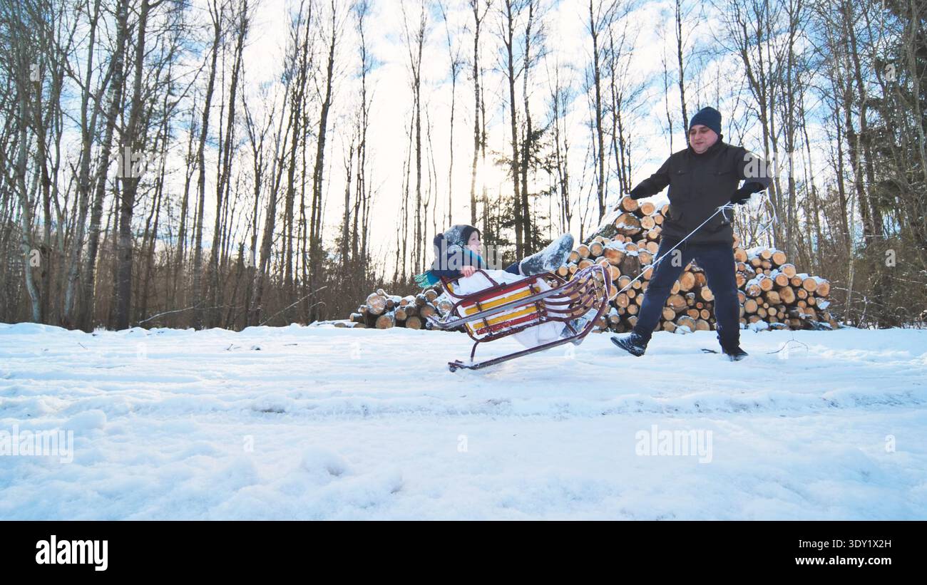 Papà tira suo figlio su una slitta in una foresta innevata accanto a una pila di legna da ardere in una giornata invernale di sole Foto Stock