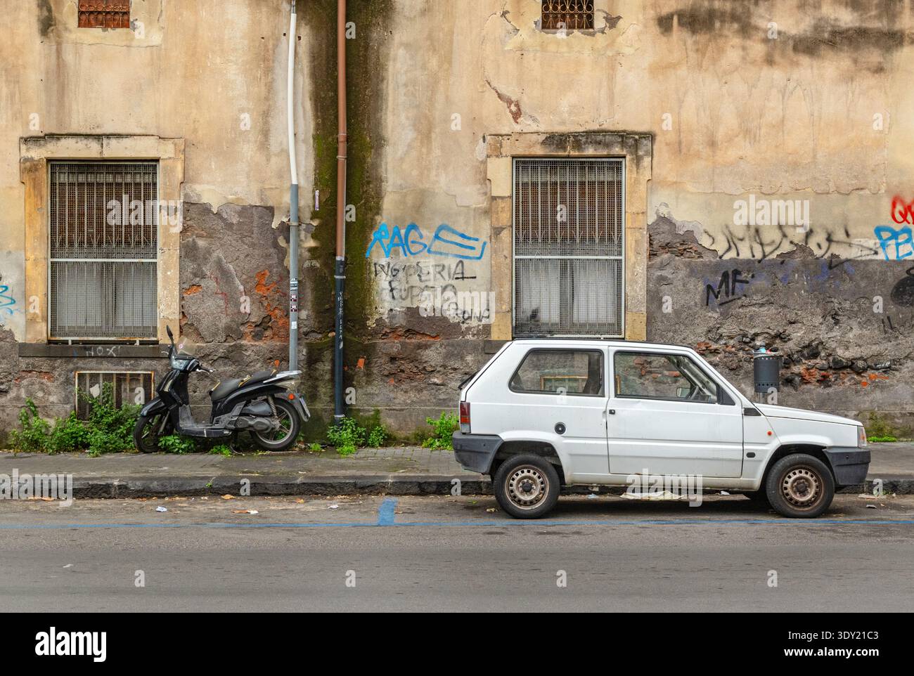 Una scena di strada all'aperto in via Plebiscito nella città di Catania, Sicilia, Italia. Il muro dell'edificio, con un'auto e uno scooter parcheggiati in strada. Foto Stock