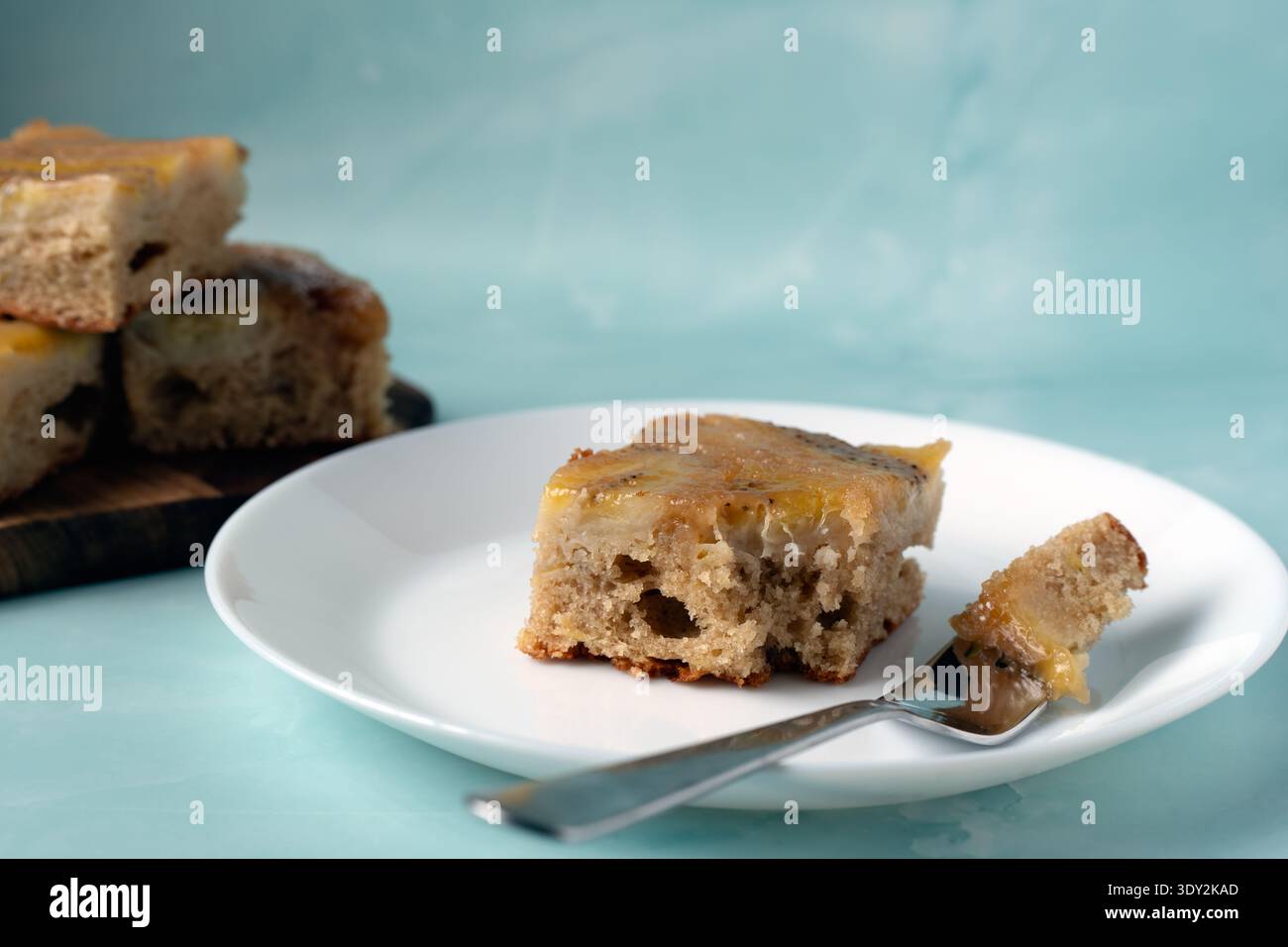 Un unico quadrato di pane alla banana umido su un piatto bianco con una forchetta. Consistenza morbida e copertura caramellata alla banana visibile Foto Stock
