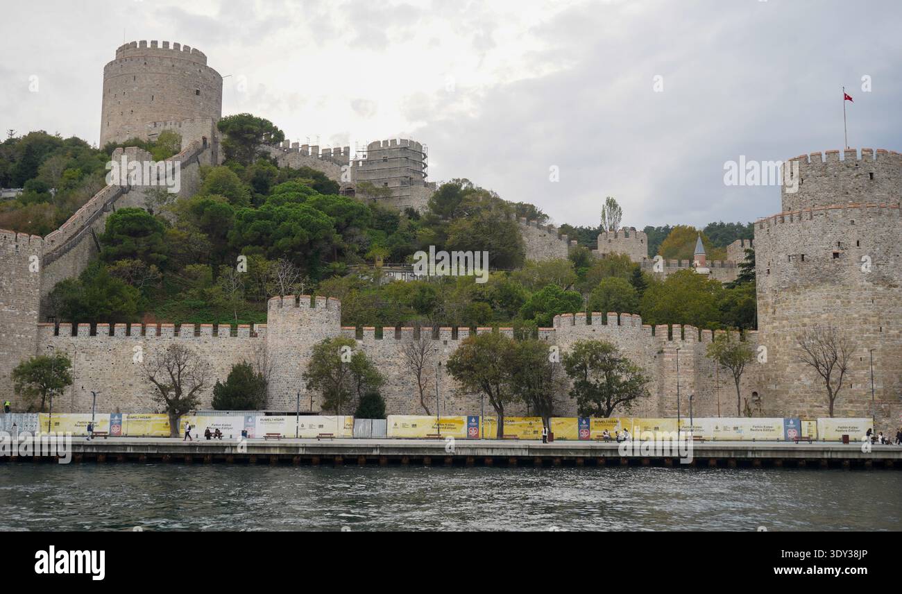 Le mura di pietra di Rumeli Hisarı si affacciano sul Bosforo. Alcuni passanti sparsi camminano lungo il tranquillo lungomare sottostante, incorniciato da fortificazioni storiche. Foto Stock