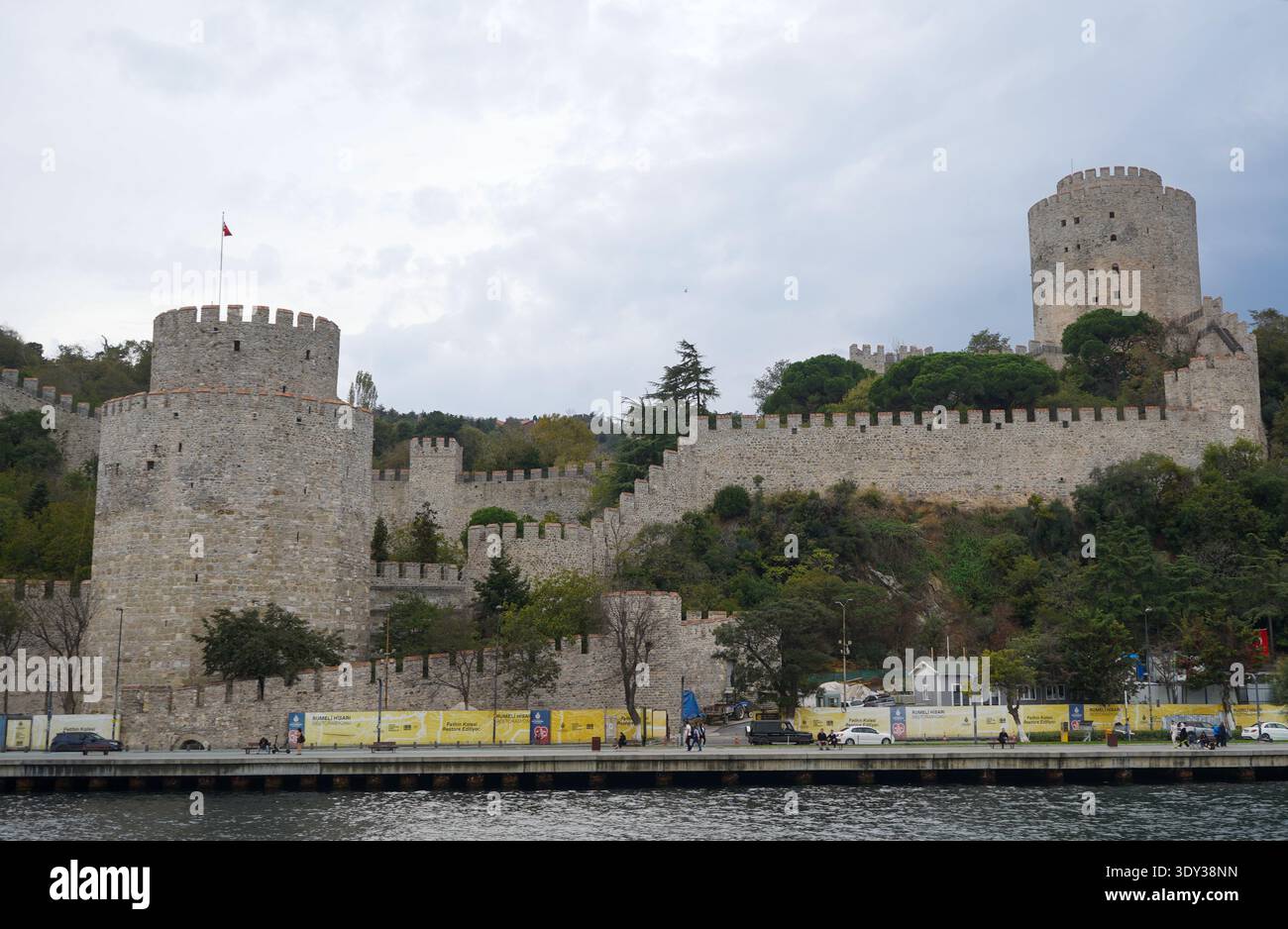 Le mura di pietra di Rumeli Hisarı si affacciano sul Bosforo. Alcuni passanti sparsi camminano lungo il tranquillo lungomare sottostante, incorniciato da fortificazioni storiche. Foto Stock
