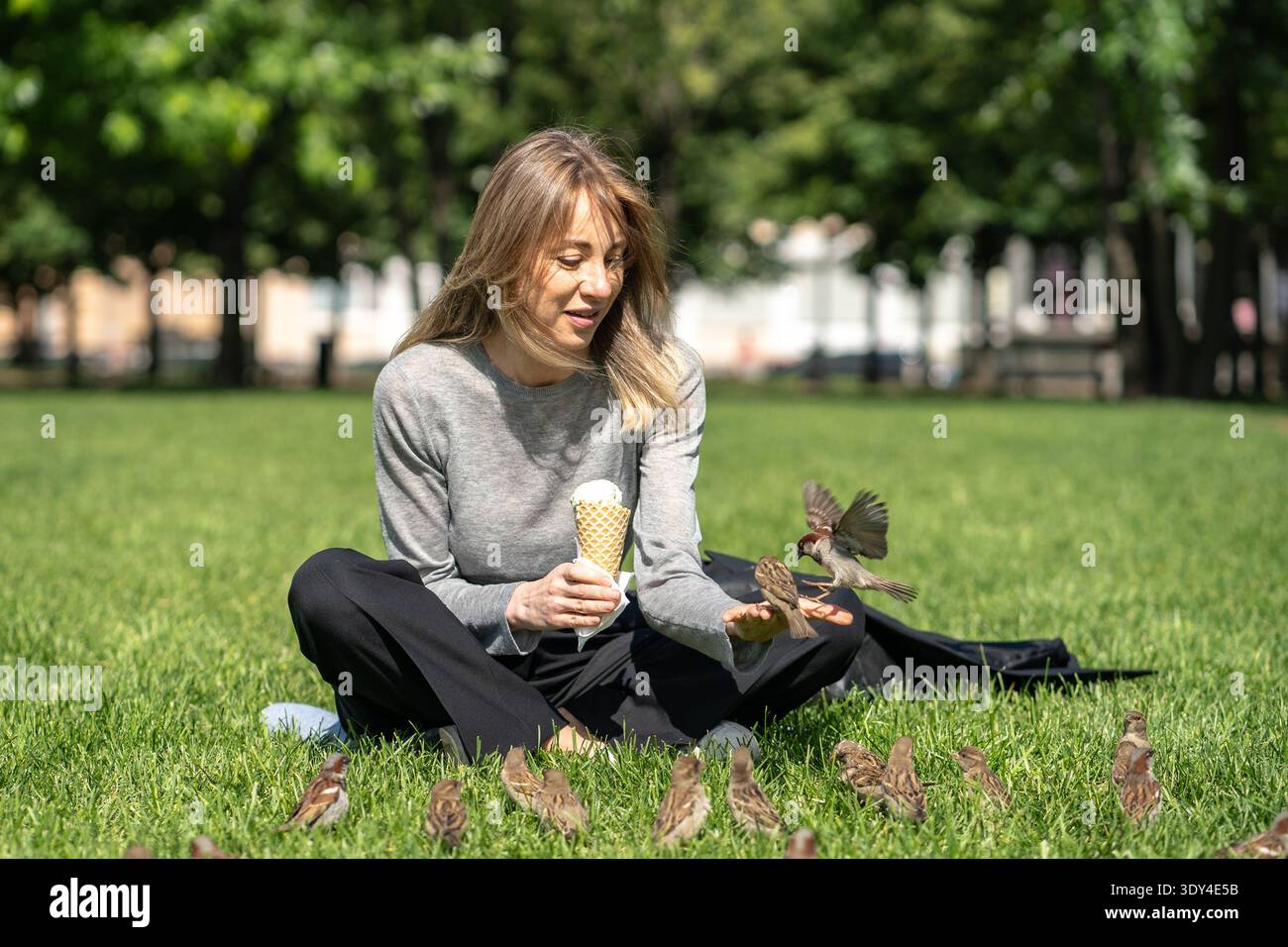 Donna d'affari positiva che si rilassa nel parco seduto sull'erba mangiando gelato e dando da mangiare agli uccelli curiosi. Foto Stock