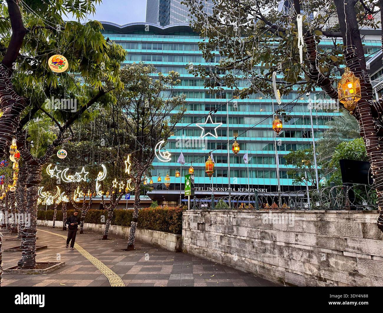 Giacarta - 5 marzo 2026 : lanterne Ramadan e decorazioni a mezzaluna sono appese agli alberi fuori dall'Hotel Indonesia Kempinski Jakarta lungo un marciapiede affollato - Immagine stock catturata con smartphone