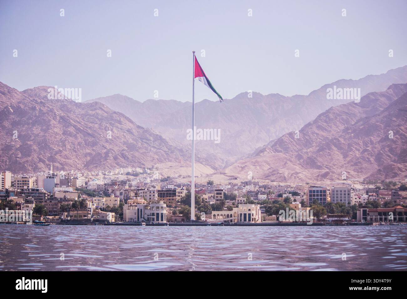L'imponente bandiera della rivolta araba che sventola dall'Aqaba Flagpole sopra lo skyline della città costiera e le aspre montagne desertiche in Giordania, viste dall'acqua. Foto Stock