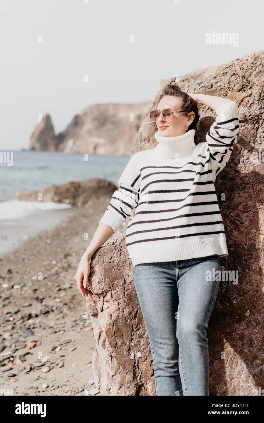 Donna spiaggia costa giovane bruna in maglione a righe si appoggia su una roccia che guarda al mare e alle scogliere lontane Foto Stock