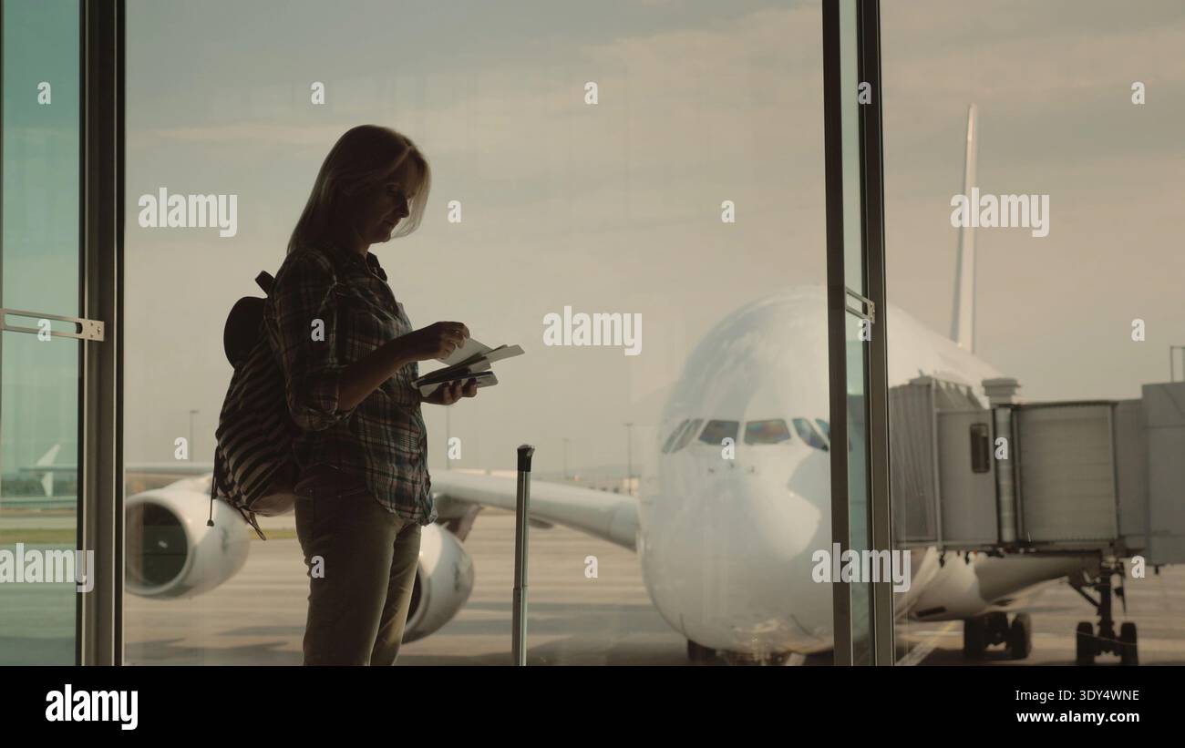 La silhouette di una donna con i documenti d'imbarco in mano, in attesa di atterrare sul suo volo. Si trova alla finestra del terminal dell'aeroporto Foto Stock