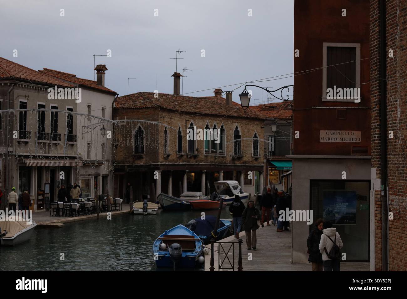Canale fiancheggiato da piccole barche ed edifici storici, dove i pedoni camminano lungo la stretta strada sul lungomare sotto un cielo coperto. Foto Stock