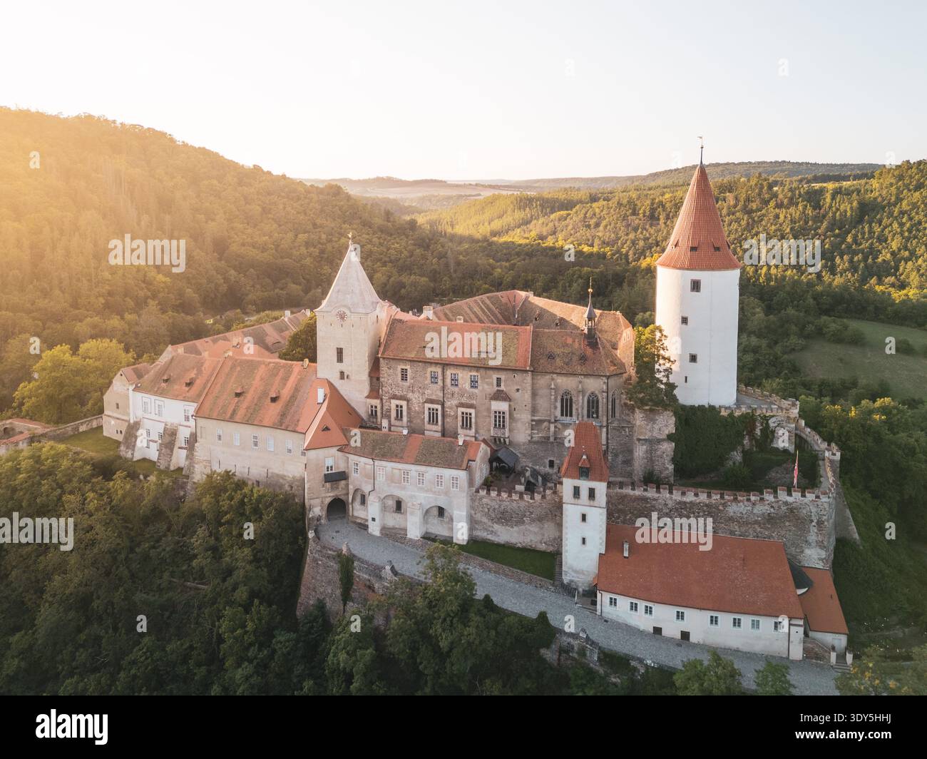 Il Castello di Krivoklat si trova in alto nella Boemia centrale, mostrando la sua architettura medievale sullo sfondo di lussureggiante vegetazione durante il tramonto, che proietta una luce dorata sul paesaggio. Foto Stock