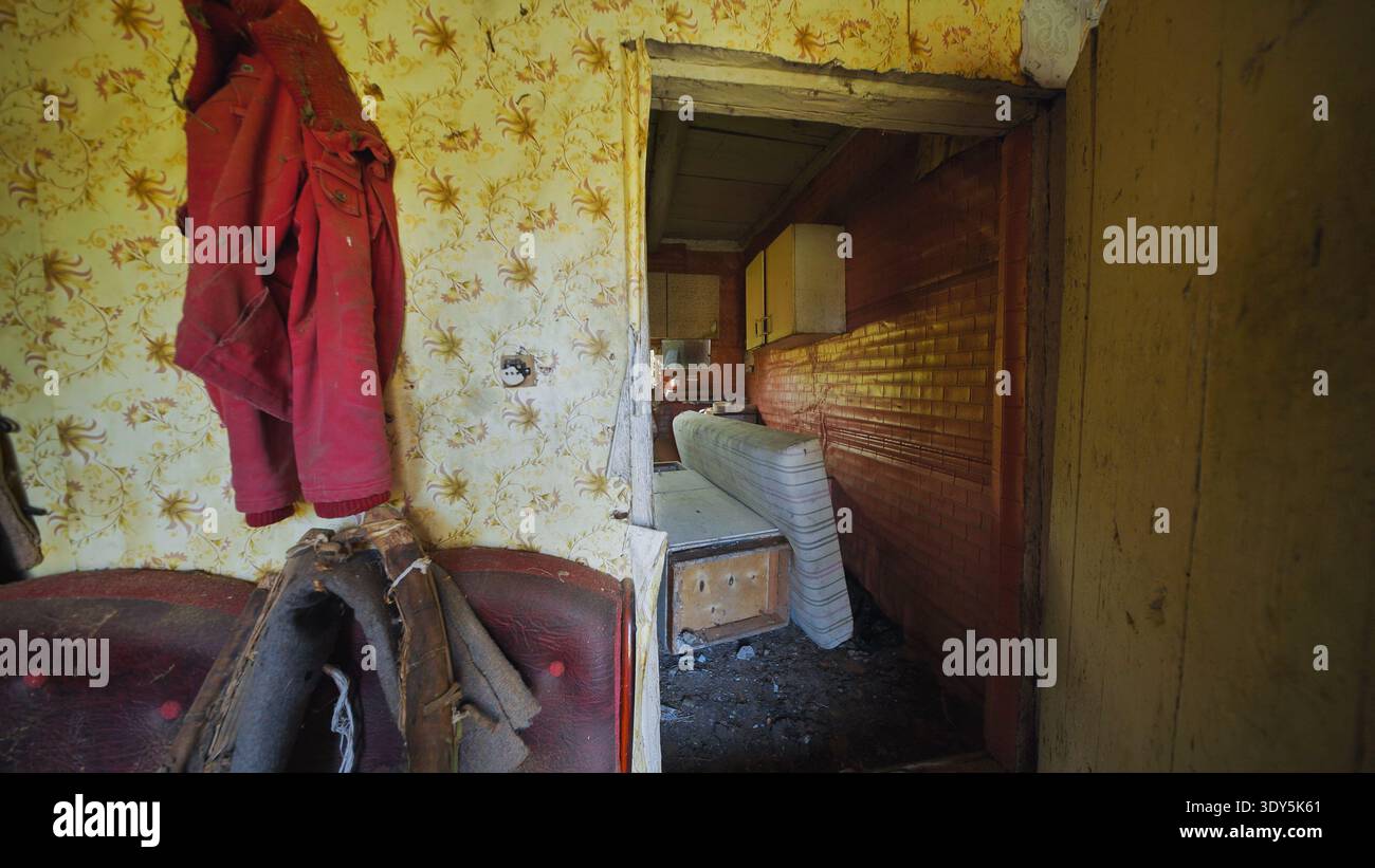 Interni di una vecchia casa abbandonati con una giacca rossa appesa su una carta da parati floreale in decomposizione Foto Stock