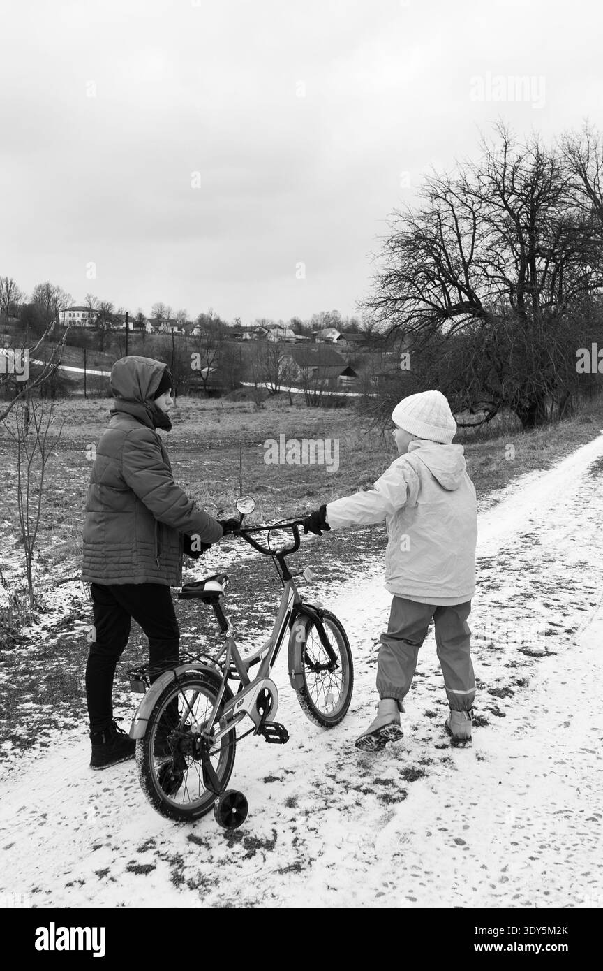 Un bambino riceve una guida su una bicicletta da un adulto su un sentiero innevato, circondato da alberi invernali e da un paesaggio tranquillo, che mette in mostra la gioia Foto Stock