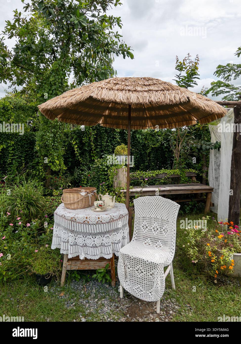 Romantico ambiente da caffe' all'aperto caratterizzato da un tavolo e una sedia ricoperti di pizzo bianco e uncinetto, in stile con un ombrello di paglia in un lussureggiante cott inglese verde Foto Stock