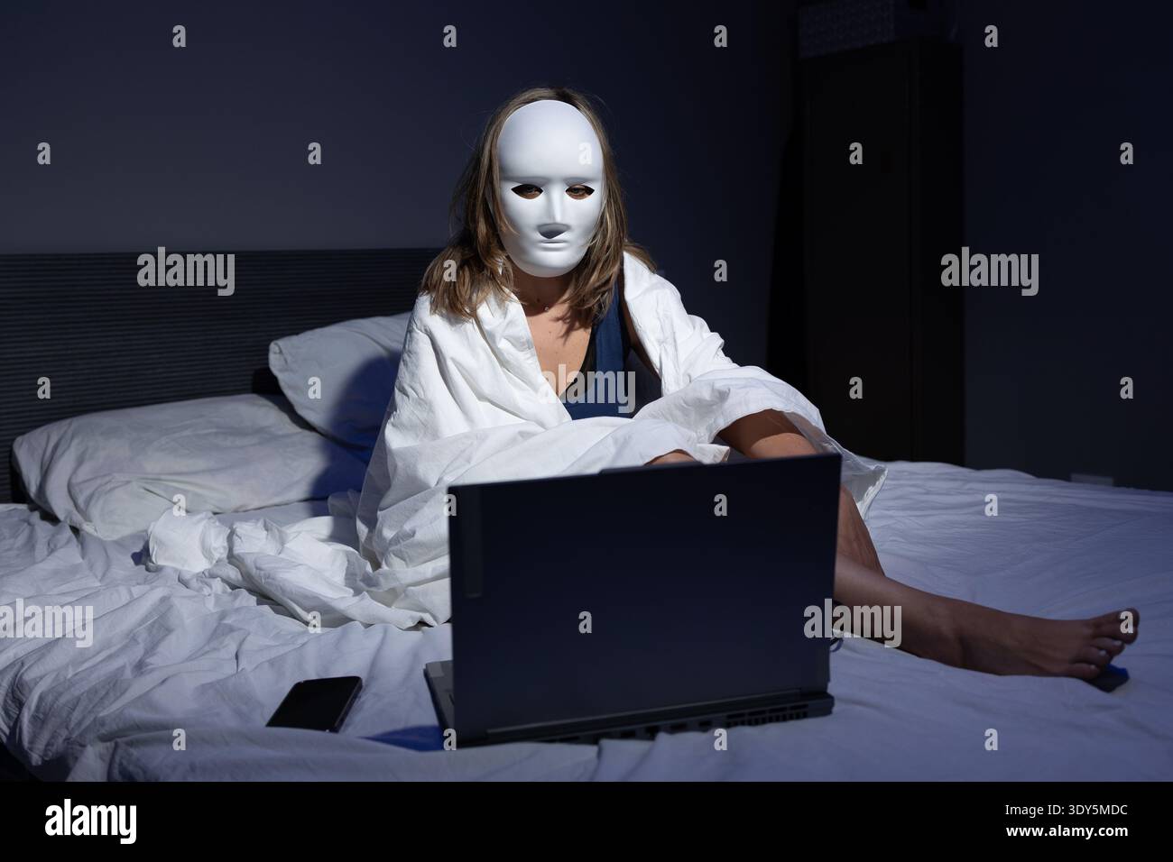 Una persona con una maschera bianca è seduta su un letto, avvolta in una coperta mentre è concentrata su un laptop. La camera oscura mette in risalto i temi della privacy, della protezione dei dati e della prevenzione delle frodi nell'era digitale. Foto Stock