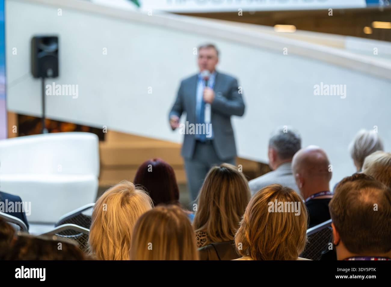 Man in Suit parla con un pubblico attento in un ambiente luminoso e moderno. Il presentatore tiene il microfono e il clicker wireless mentre la folla ascolta durante l'evento. Foto Stock