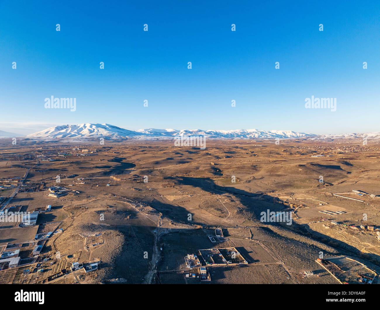 Vista aerea delle aride pianure desertiche sotto il cielo azzurro. Le montagne innevate si innalzano in lontananza, generando lunghe ombre. Edifici e strade sparse Foto Stock