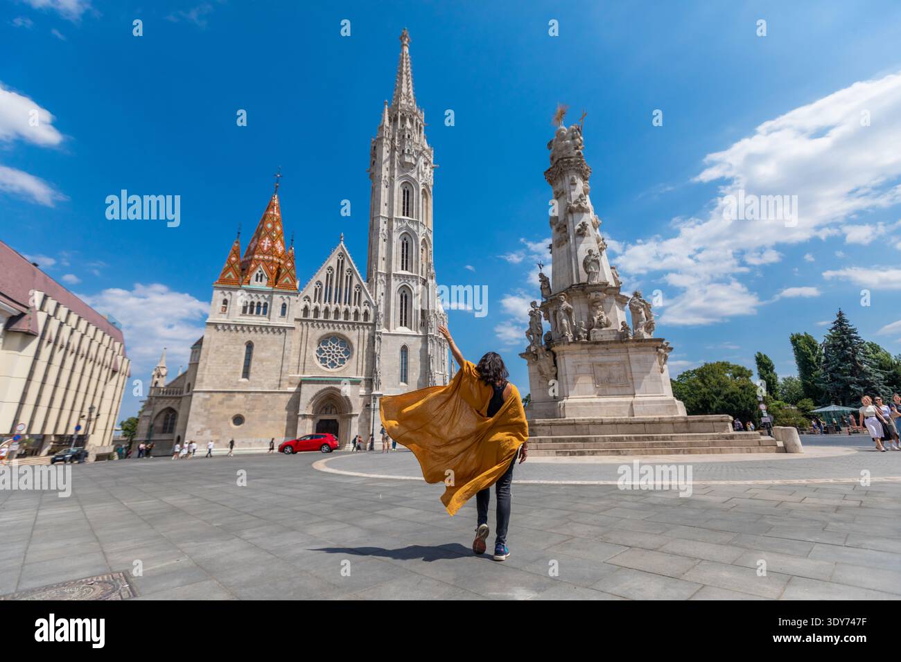 Il viaggiatore con il mantello giallo cammina verso la chiesa di Mattia e la colonna della Santissima Trinità a Budapest in una giornata di sole Foto Stock