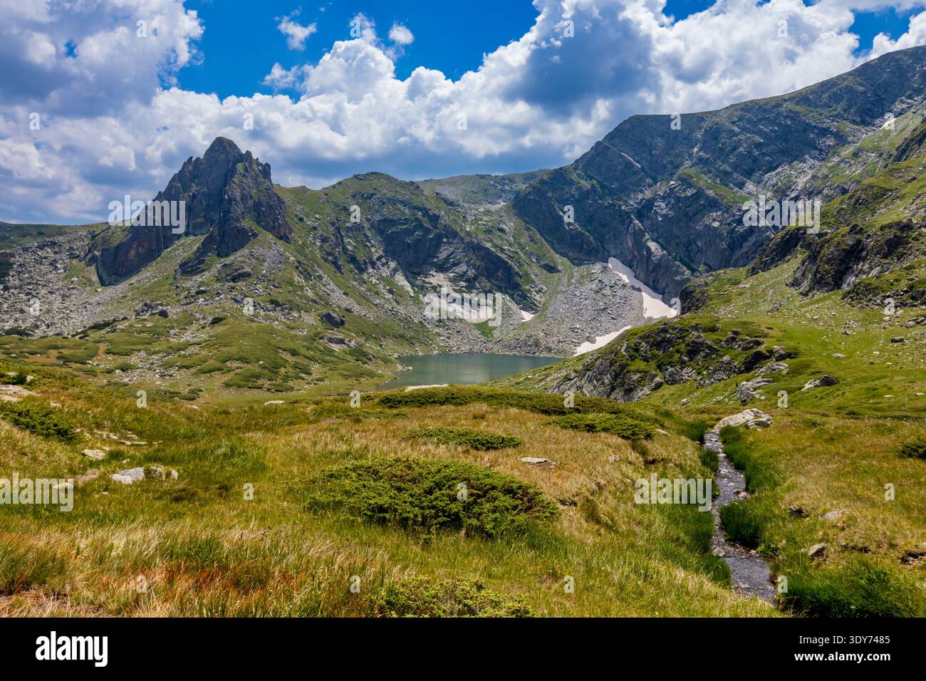 Il prato e il torrente scorrono sotto le vette delle montagne con un lago in lontananza nei Monti Rila Bulgaria Foto Stock