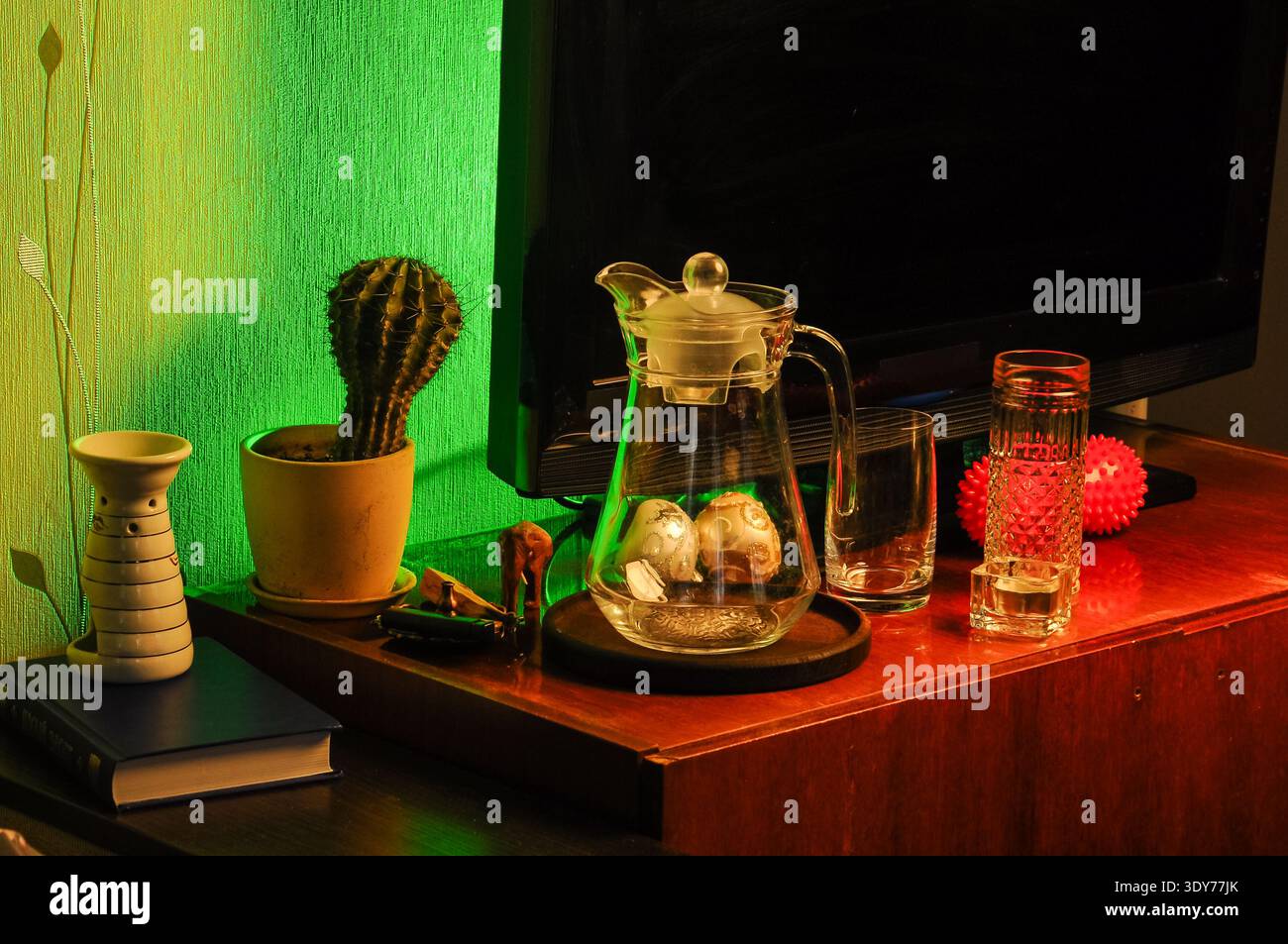 Una collezione di oggetti per la casa, tra cui un cactus in vaso, una caraffa di vetro con sfere decorative, bicchieri vuoti e una palla rossa sono disposti su una Foto Stock