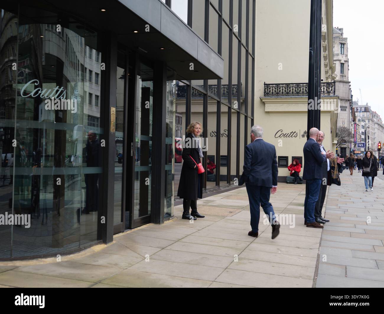 Professionisti ben vestiti e adatti si riuniscono fuori dalla prestigiosa sede di Coutts & Co. Al 440 di The Strand, Londra, mentre una donna siede sulla sua valigia a fare una sigaretta arrotolata direttamente sotto lo storico emblema delle tre corone della banca. Catturata nel febbraio 2026, questa scena presenta un'impressionante giustapposizione del divario socio-economico nella capitale britannica. Il mondo del private banking, che richiede ai clienti di detenere almeno 1 milione di sterline in asset investibili, incontra la realtà della vita di strada transitoria in una delle strade più trafficate di Londra Foto Stock